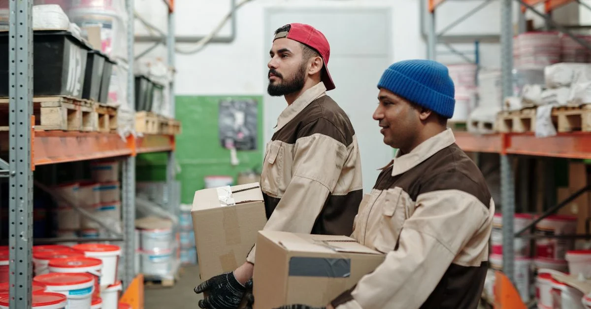 Dos trabajadores en un almacén llevando cajas, estantes organizados al fondo.
