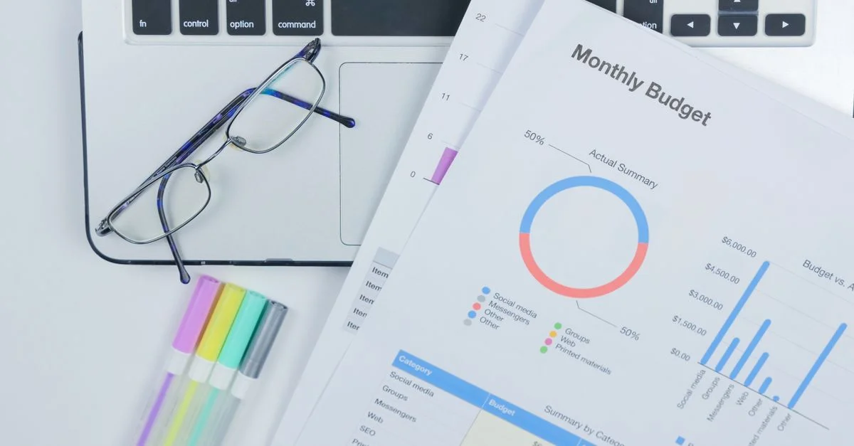 Un escritorio de oficina limpio que presenta un portátil, gafas y un colorido informe presupuestario.