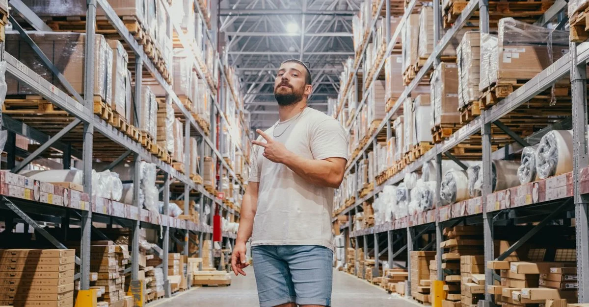 Joven posando en un pasillo de almacén rodeado de estanterías y paquetes dentro de una tienda espaciosa.